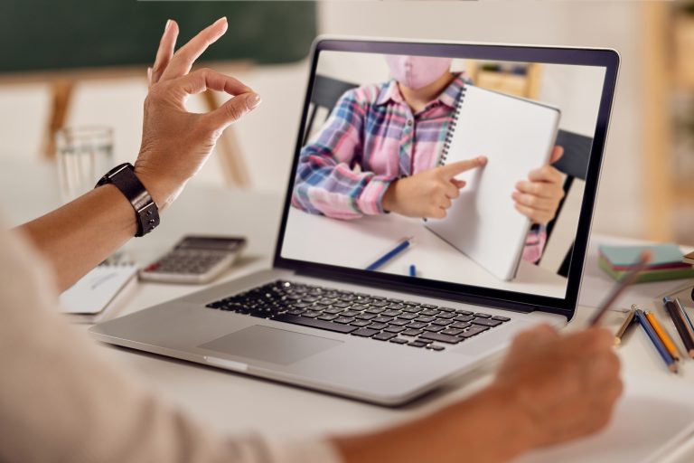 Dziecko w maseczce pokazujące pustą kartkę w zeszycie podczas lekcji online na ekranie laptopa. Nauczycielka odpowiada gestem „OK”, siedząc przy biurku.